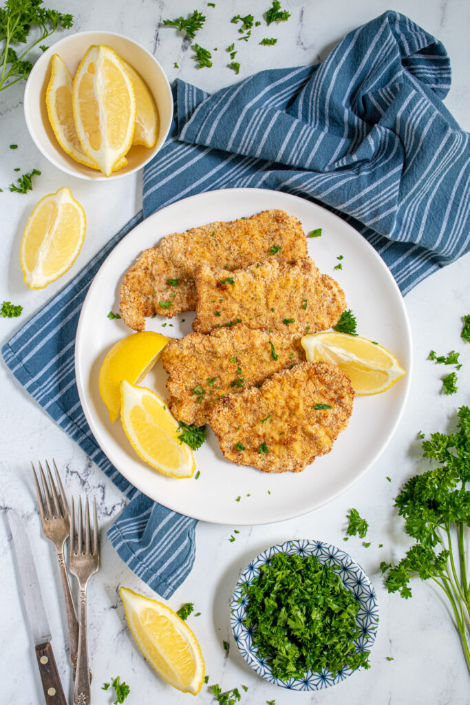 Overhead view of a tabletop with a white plate full of air fryer pork schnitzel garnished with fresh parsley and surrounded by lemon wedges.