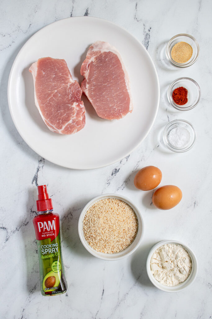 Ingredients for sir fryer schnitzel laid out on a tabletop including pork chops, breadcrumbs, flour, and eggs.