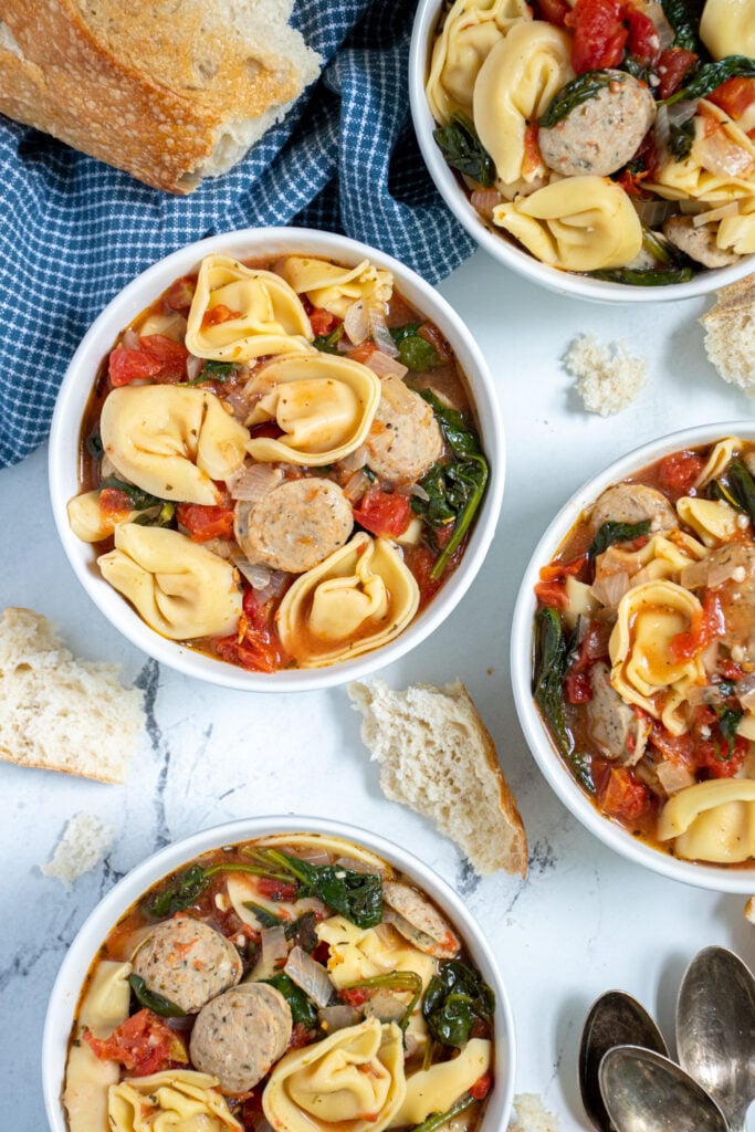 Overhead view of a few white bowls of chicken sausage tortellini soup with spinach and diced tomatoes.