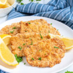 Pinterest Pin with text 'Air Fryer Pork Schnitzel', image of a white plate full of golden brown schnitzel garnished with parsley and lemon wedges.