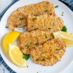 Pinterest Pin with text 'Air Fryer Pork Schnitzel', image of a white plate full of golden brown schnitzel garnished with parsley and lemon wedges.