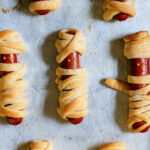 Pinterest Pin with text 'Halloween Mummy Dogs', image of mummy dogs with mustard eyes laid out on a sheet pan.