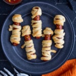 Pinterest Pin with text 'Halloween Mummy Dogs', image of mummy dogs with mustard eyes on a black plate.