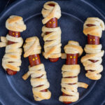 Pinterest Pin with text 'Halloween Mummy Dogs', image of mummy dogs with mustard eyes on a black plate.