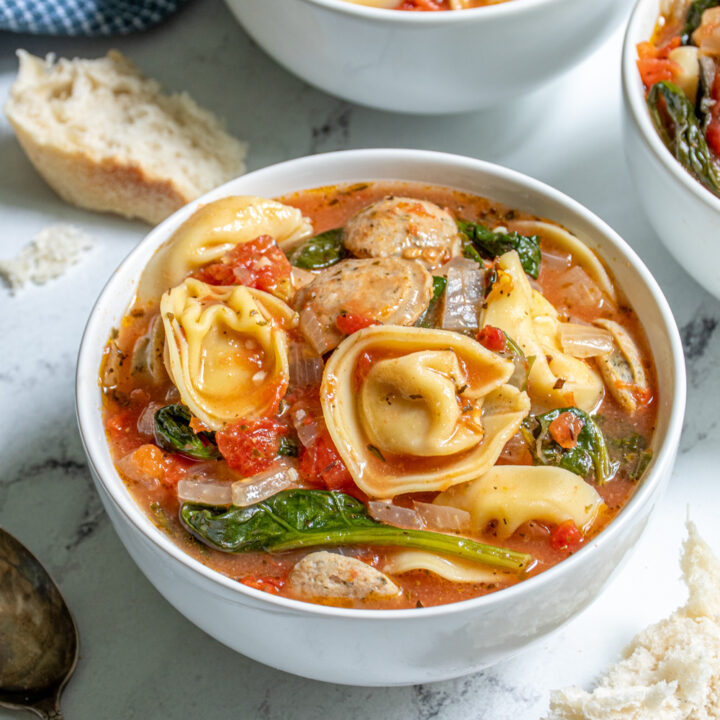 Close up of a white bowl filled with tortellini soup with chicken sausage, spinach, and diced tomatoes.