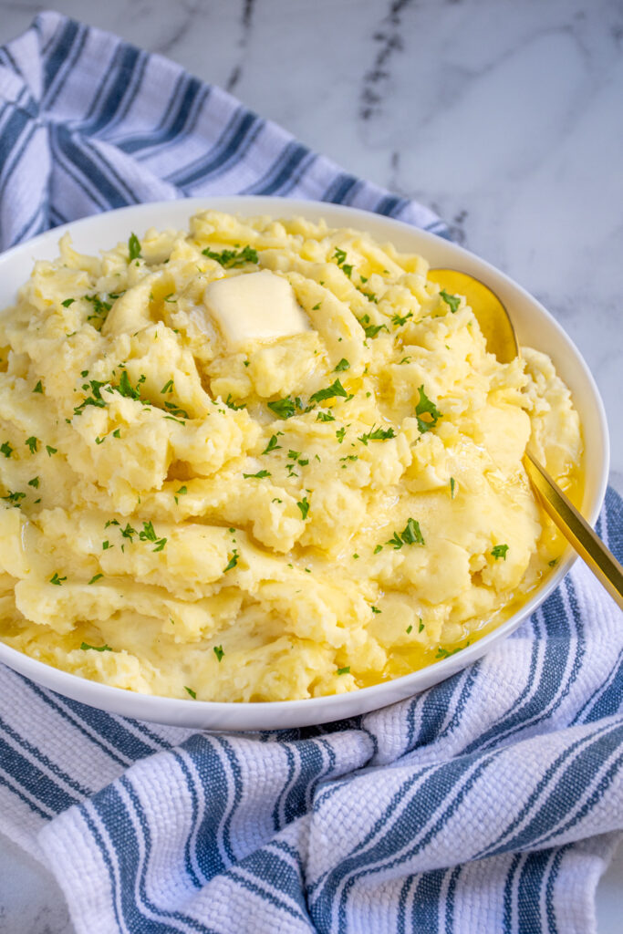 White bowl on a blue striped towel filled with mashed potatoes and topped with melting butter and garnished with fresh parsley.