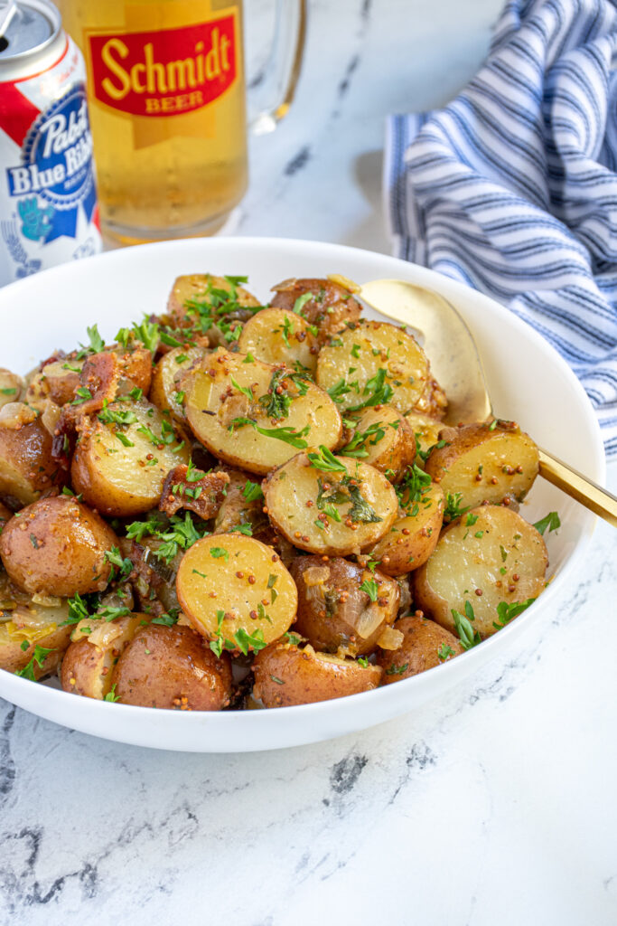 Side view of a large white bowl filled with German potato salad covered in a mustard sauce, with bacon bits and fresh parsley.