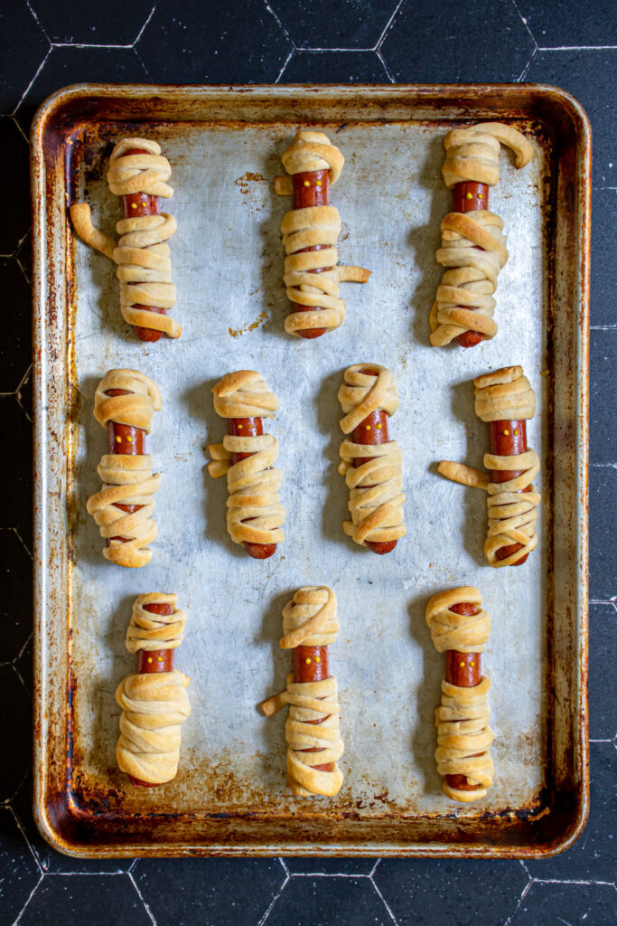 Sheet pan full of mummy hot dogs cooked golden brown with little mustard dots for mummy eyes.