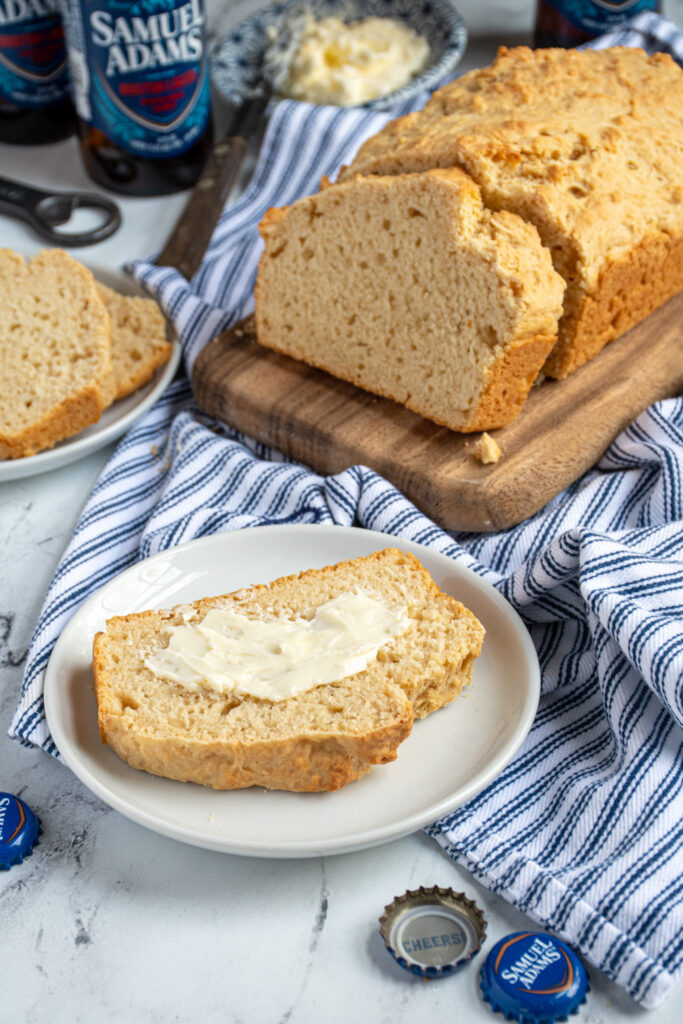A slice of buttered beer bread on a plate sitting next to the full loaf.