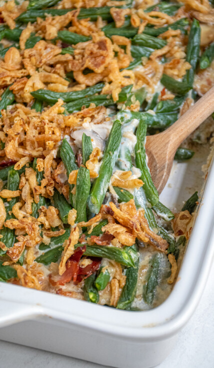 A white baking dish with green bean casserole with bacon with a serving spoon scooping some out.
