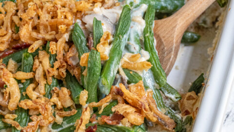 A white baking dish with green bean casserole with bacon with a serving spoon scooping some out.