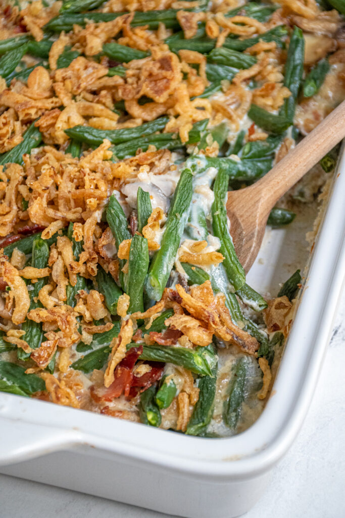 A white baking dish with green bean casserole with bacon with a serving spoon scooping some out.