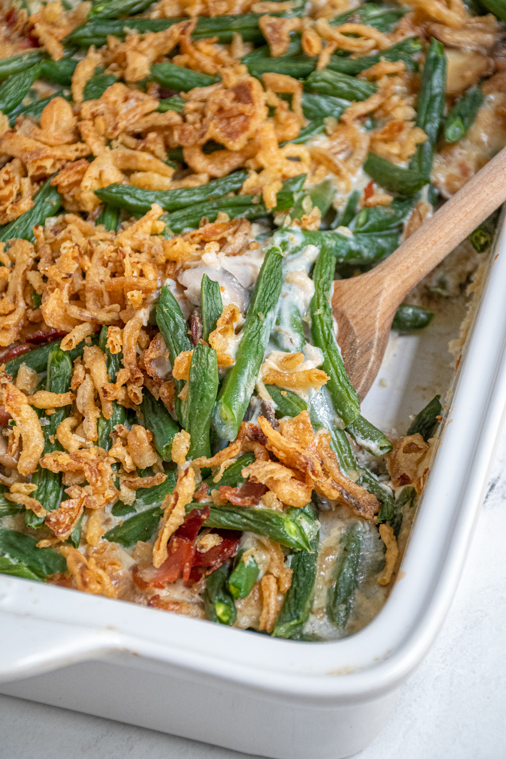 A white baking dish with green bean casserole with bacon with a serving spoon scooping some out.