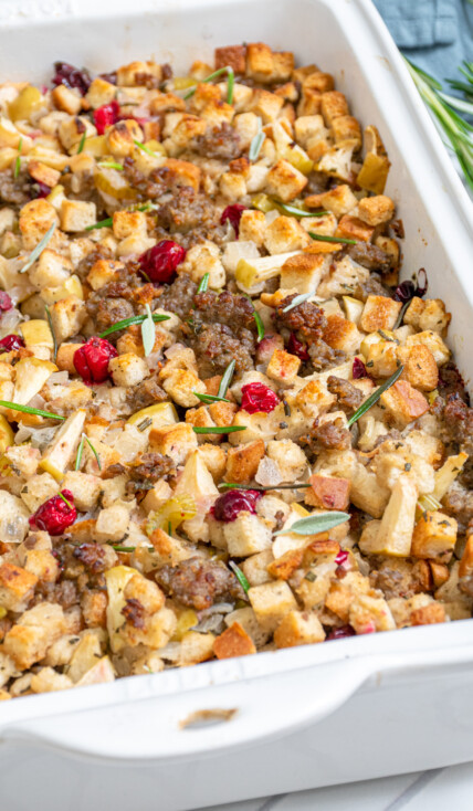 A white casserole dish with Sausage Cranberry Apple Stuffing with pops od red cranberries and garnished with fresh rosemary.