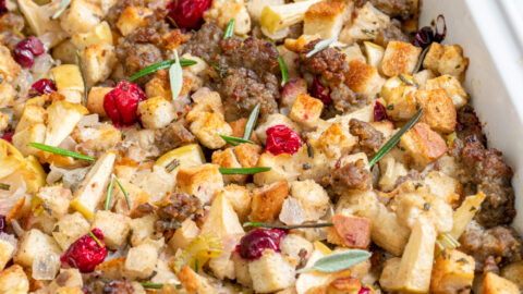A white casserole dish with Sausage Cranberry Apple Stuffing with pops od red cranberries and garnished with fresh rosemary.