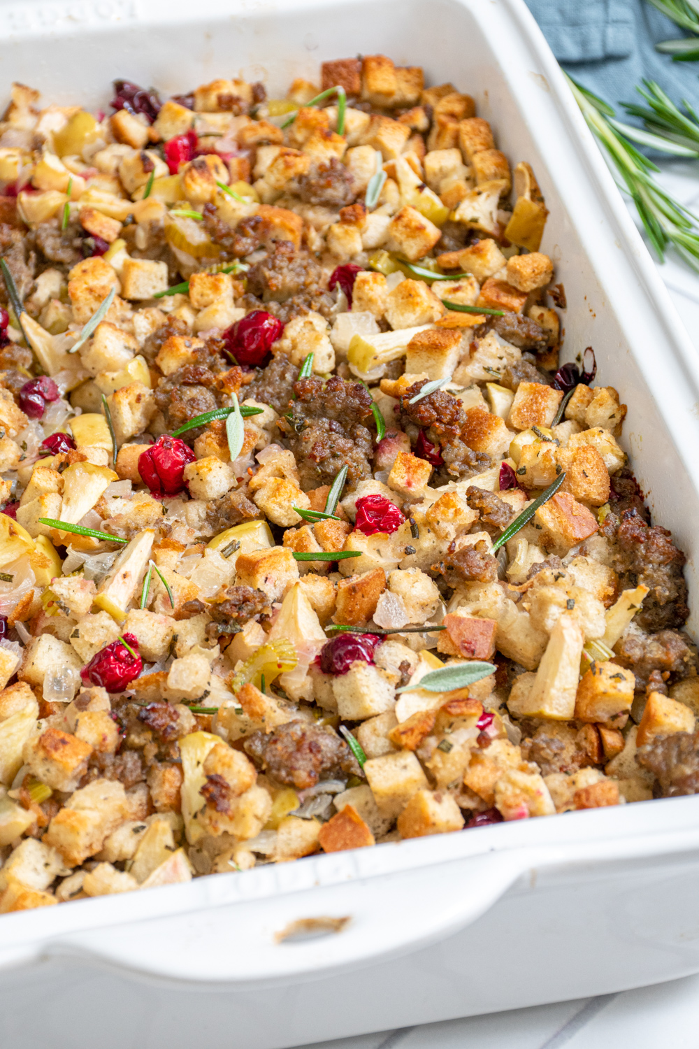 A white casserole dish with Sausage Cranberry Apple Stuffing with pops od red cranberries and garnished with fresh rosemary.