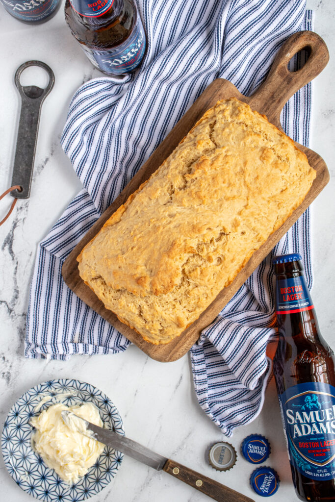 A loaf of beer bread on a cutting board surrounded by beer bottles and a bowl of butter.