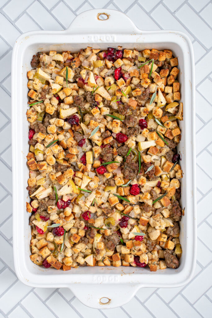 Overhead of casserole dish of stuffing with cranberries, apples, and sausage garnished with fresh rosemary.