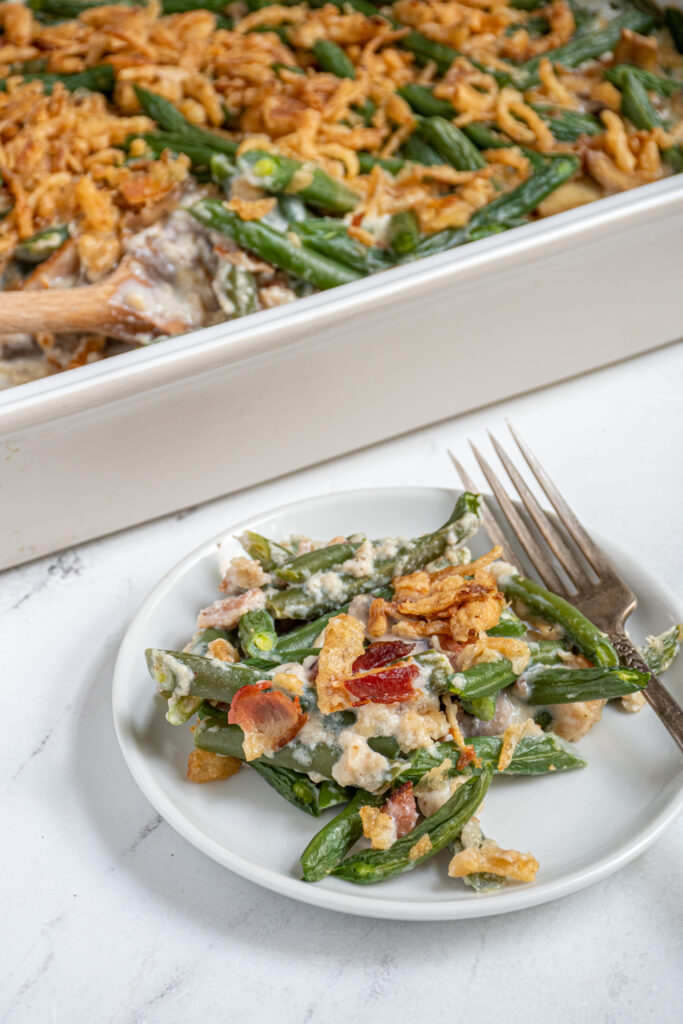 A small plate with a scoop of green bean casserole with bacon next to a casserole dish.