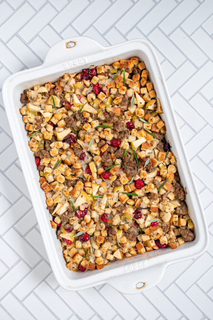 Overhead view of a white casserole dish with stuffing with sausage, cranberries, and apples garnished with fresh rosemary.
