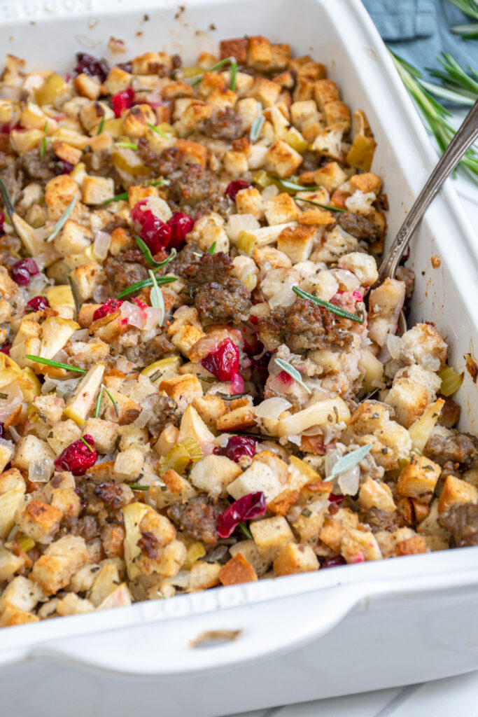 Close up view of a spoon scooping up cranberry apple sausage stuffing out of a white casserole dish.