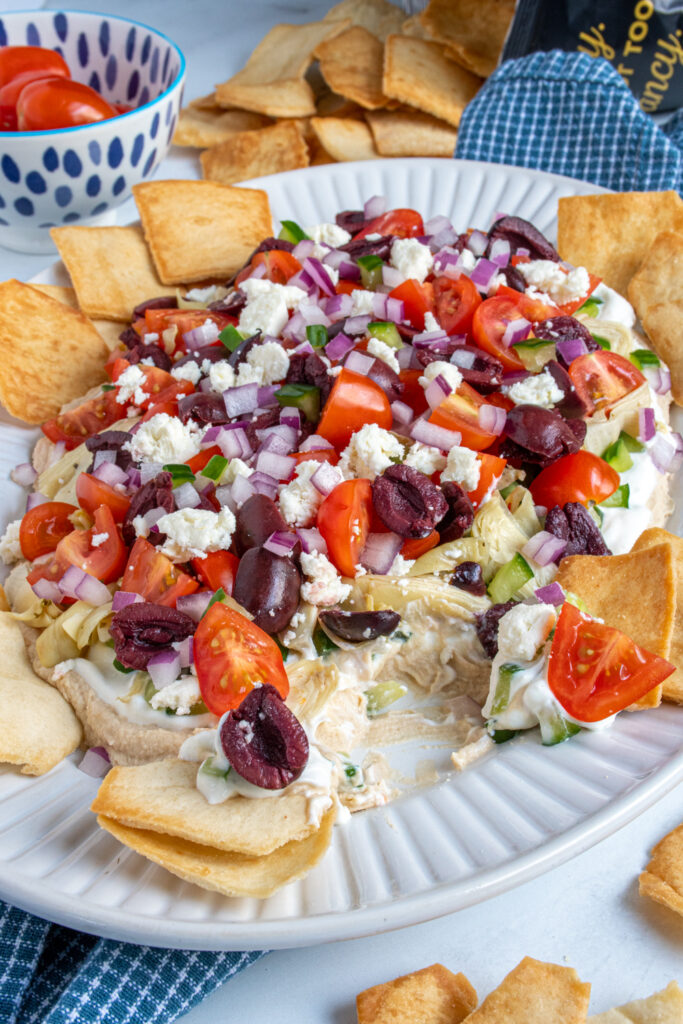 Close up of a platter layered with greek dip with a pita chip scooping some dip out.