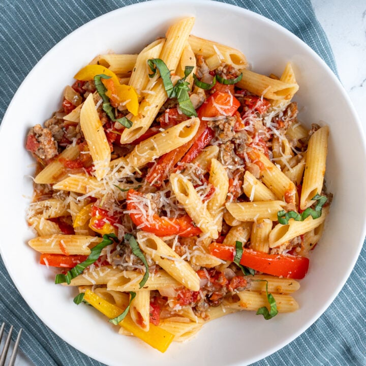An overhead view of a white bowl filled with penne pasta, sausage, and bell peppers and garnished with parmesan cheese.