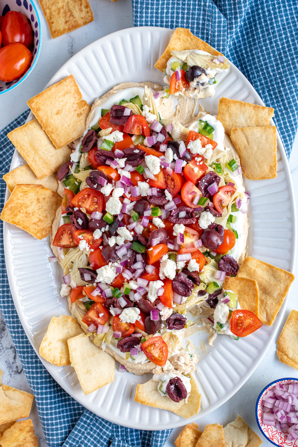 Overview of layered greek dip on a white platter showing bright tomatoes, olives, red icons, and cucumbers with pita chips scooping dip off the sides.