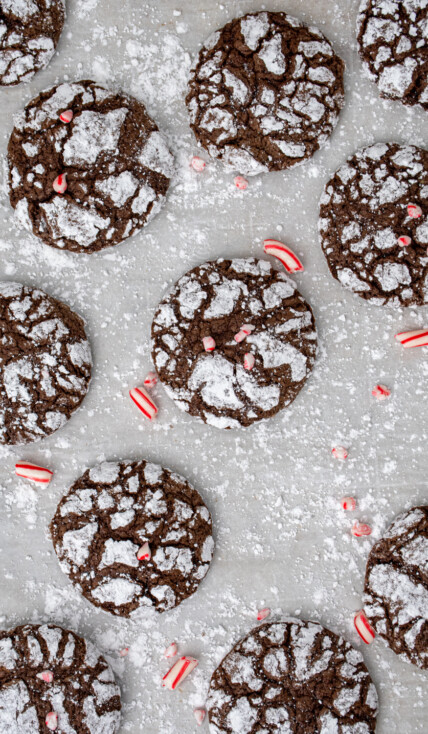 A bunch of chocolate peppermint crinkle cookies spread out over powdered sugar covered parchment paper with bits of crushed candy canes as garnish.
