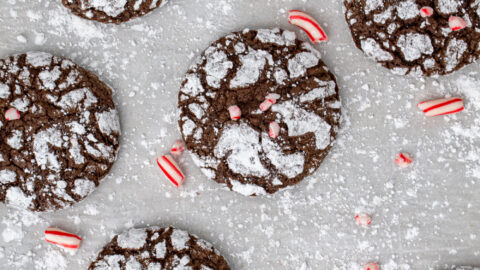 A bunch of chocolate peppermint crinkle cookies spread out over powdered sugar covered parchment paper with bits of crushed candy canes as garnish.