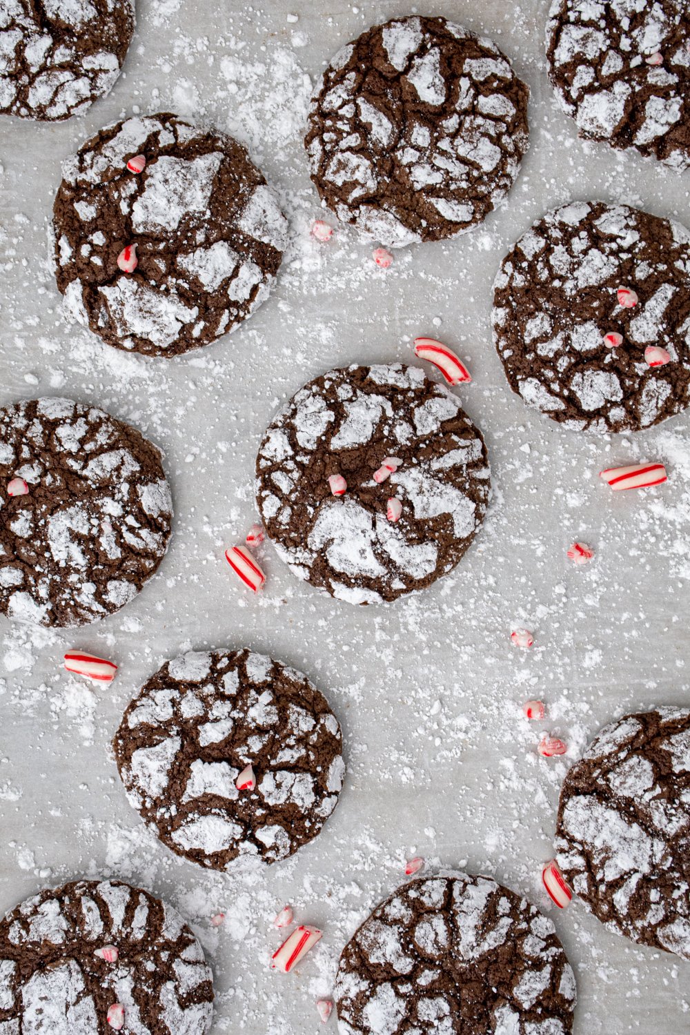 A bunch of chocolate peppermint crinkle cookies spread out over powdered sugar covered parchment paper with bits of crushed candy canes as garnish.