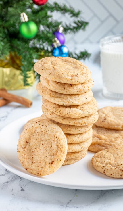 A large stack of eggnog snickerdoodles on a white plate with one eating up against the stack showing off a sugary coating filled with cinnamon and nutmeg.