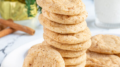 A large stack of eggnog snickerdoodles on a white plate with one eating up against the stack showing off a sugary coating filled with cinnamon and nutmeg.