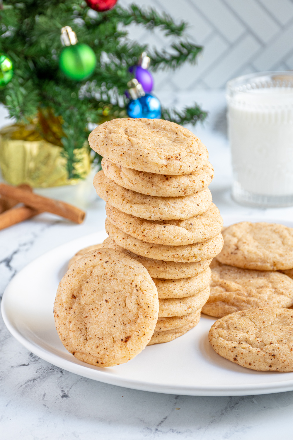 A large stack of eggnog snickerdoodles on a white plate with one eating up against the stack showing off a sugary coating filled with cinnamon and nutmeg.