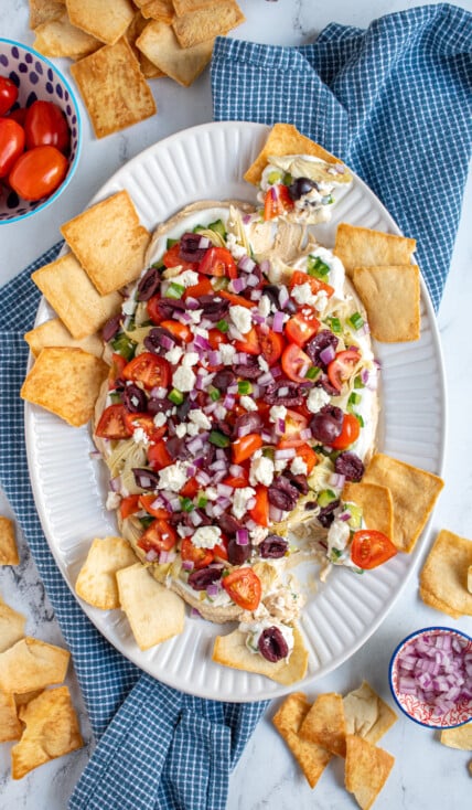 White platter full of greek layer dip with pita chips scooping the dip off of the platter.