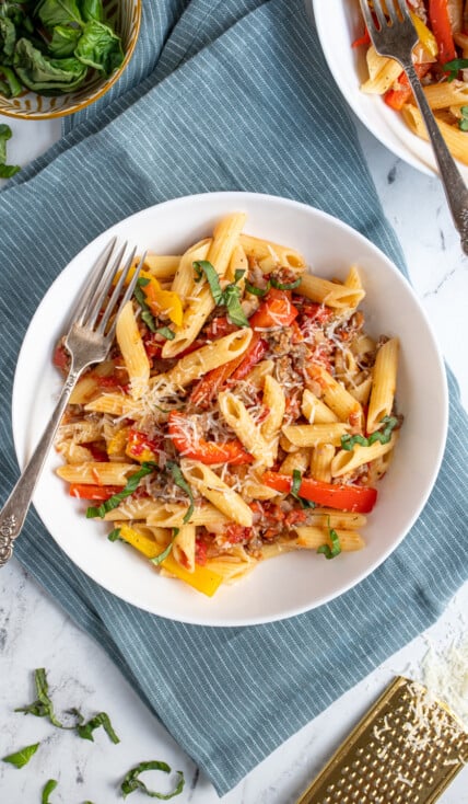Overhead view of a white bowl with a fork filled with sausage and pepper penne pasta, garnished with parmesan cheese.