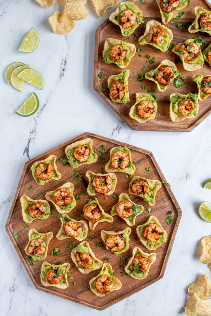An over head view of two brown platter filled with shrimp guacamole bites garnished with cilantro and lime wedges.