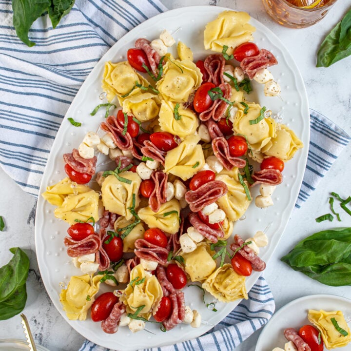 A platter surrounded by plates and fresh basil filled with tortellini skewers.