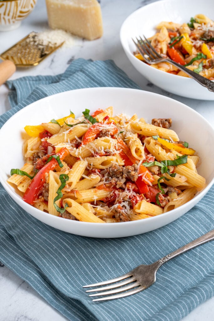A large white bowl on a blue towel filled with sausage and pepper pasta garnished with parmesan cheese and fresh basil.
