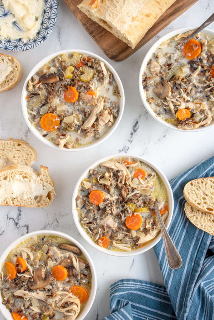 Tabletop filled with four bowls filled with slow cooker chicken wild rice soup sitting next to spoons, a blue kitchen towel, and a loaf of bread.
