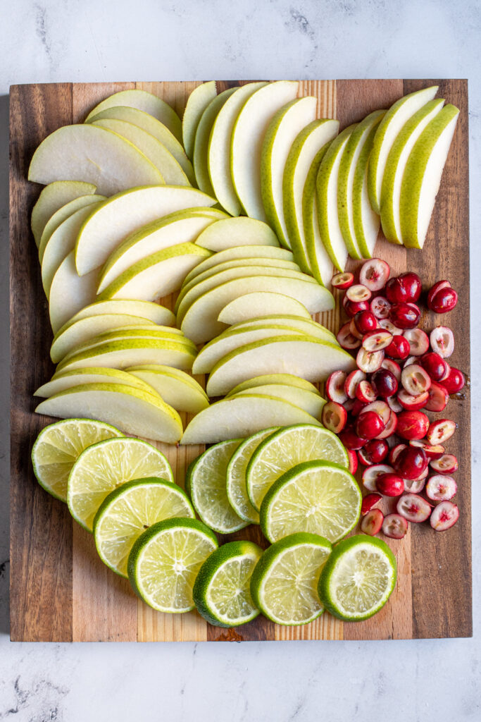 A cutting board with sliced fruit with pears, limes, and halved cranberries.