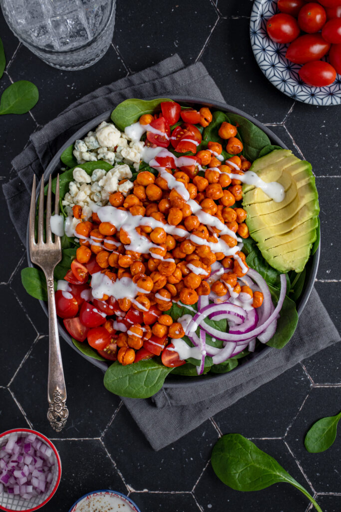 Black colored bowl filled with spinach and crispy roasted buffalo chickpeas with blue cheese, tomatoes, avocados, red onions, and a ranch drizzle.