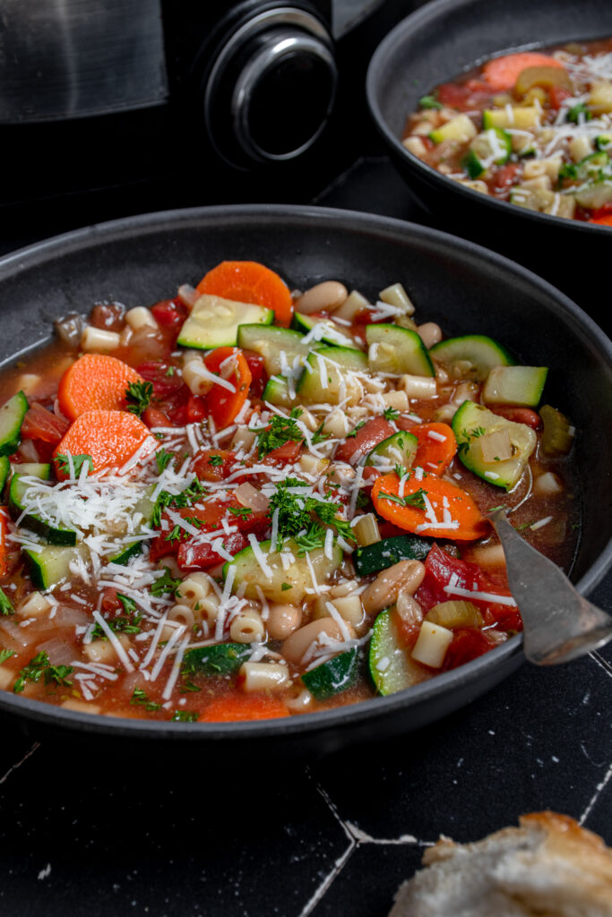 Close up of a black bowl with minestrone soup garnished with parmesan cheese and parsley.
