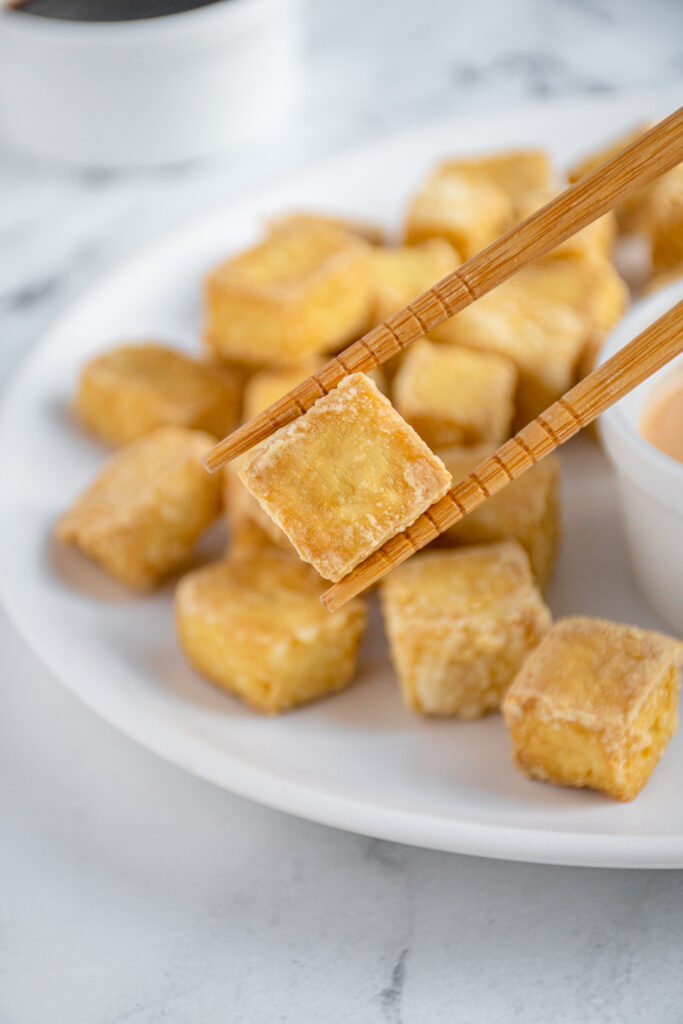 A pair of chopsticks holding a single piece of crispy tofu up close.