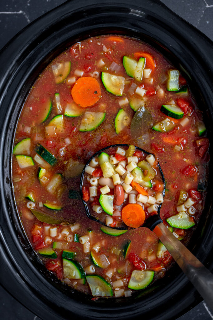 Crockpot with a ladle of minestrone soup showing off vegetables, beans, and pasta.