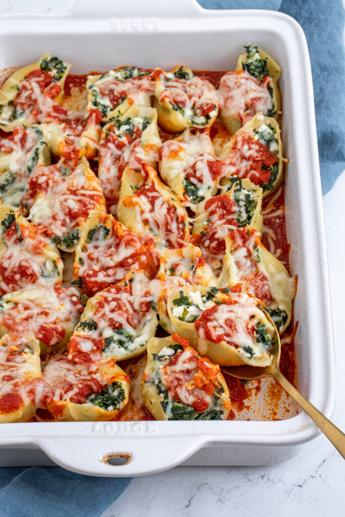 A white casserole dish filled with saucy and cheesy spinach stuffed shells with a spoon scooping one out.