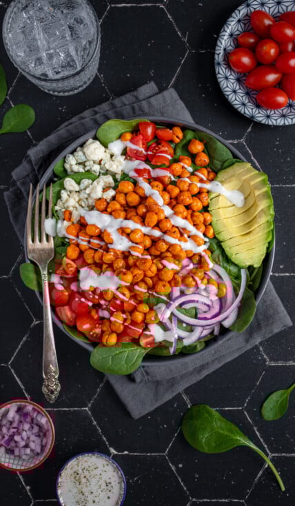 Overhead view of a black bowl on a black background, the bow is filled with buffalo chickpea salad, garnished with a ranch drizzle, avocado, red onion, and tomatoes.