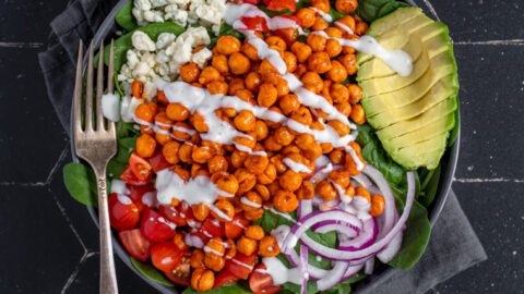 Overhead view of a black bowl on a black background, the bow is filled with buffalo chickpea salad, garnished with a ranch drizzle, avocado, red onion, and tomatoes.