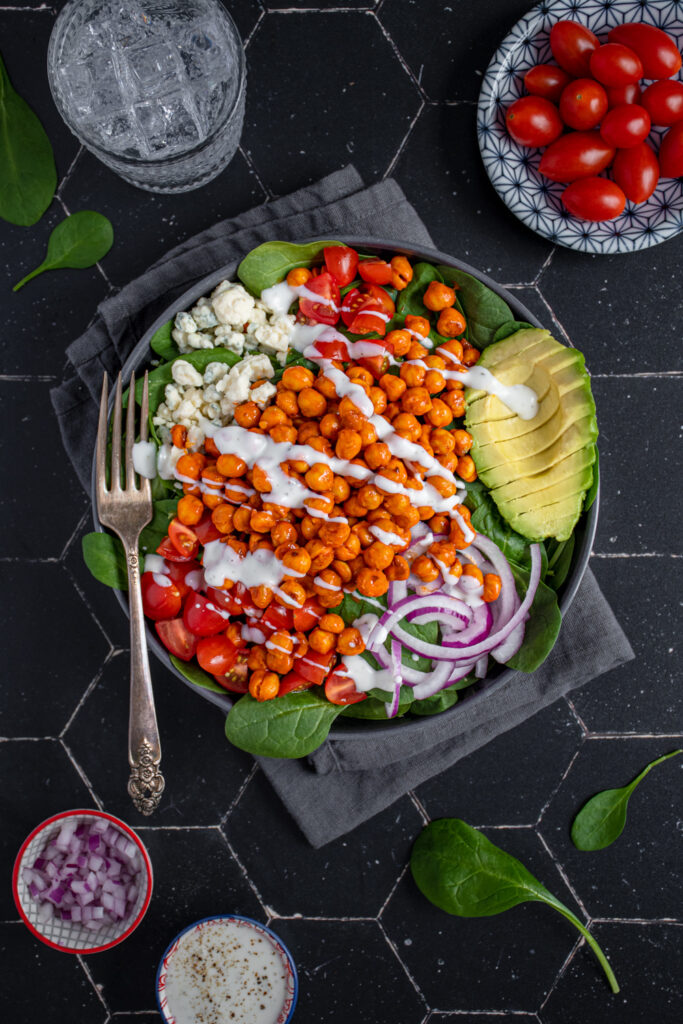 Overhead view of a black bowl on a black background, the bow is filled with buffalo chickpea salad, garnished with a ranch drizzle, avocado, red onion, and tomatoes.