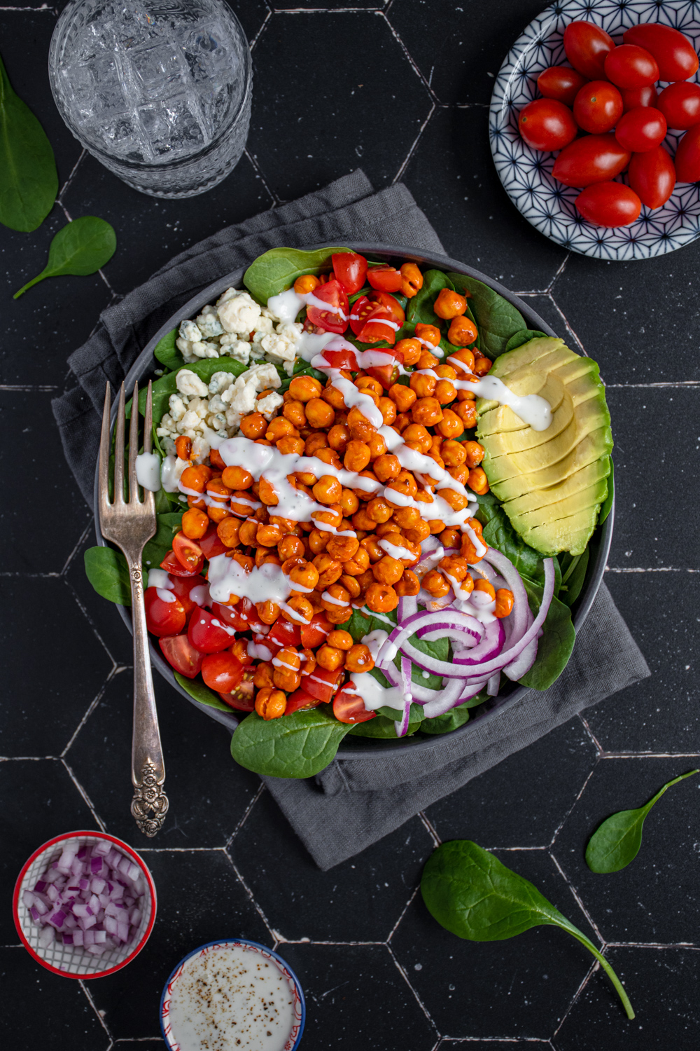 Overhead view of a black bowl on a black background, the bow is filled with buffalo chickpea salad, garnished with a ranch drizzle, avocado, red onion, and tomatoes.